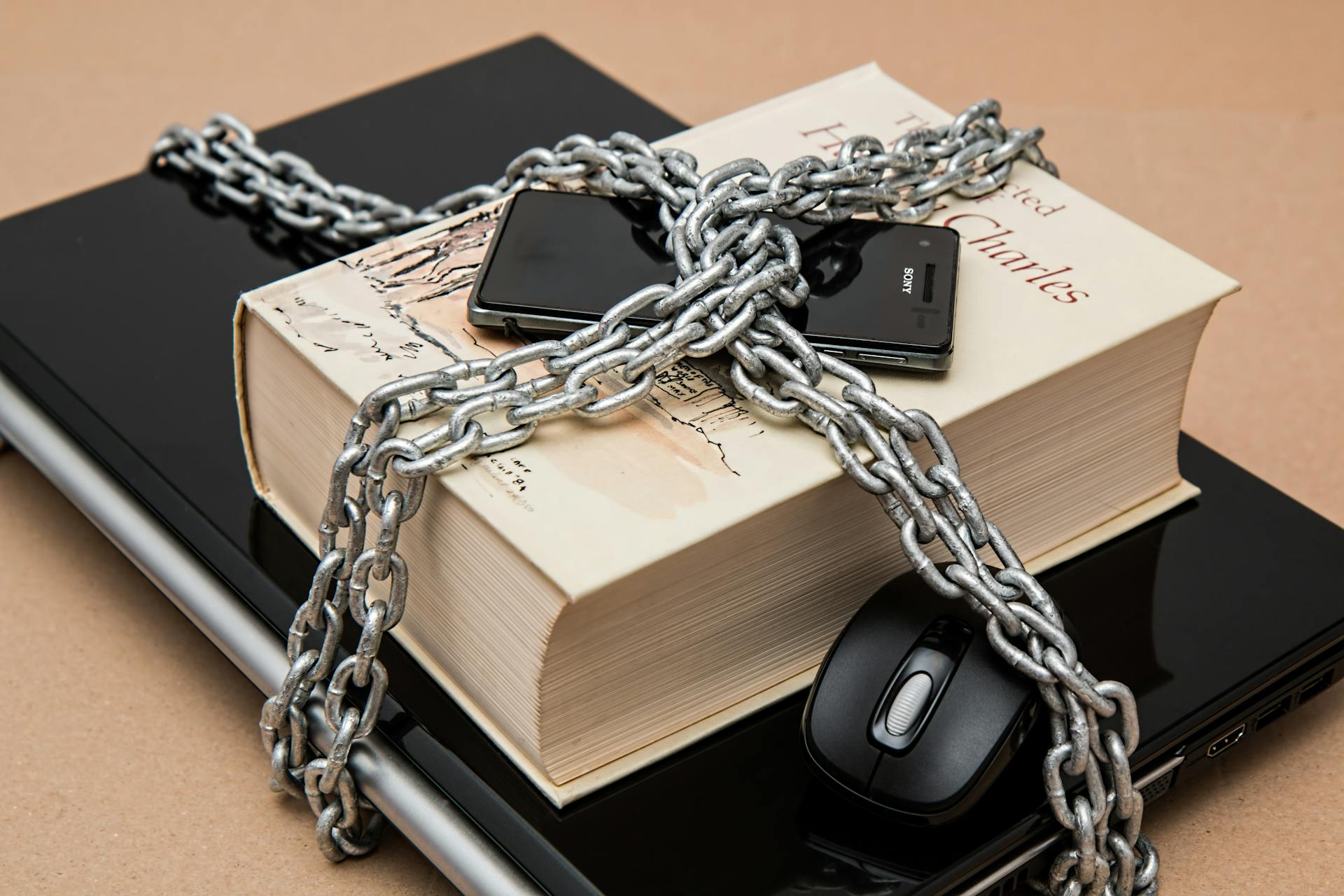 A laptop, mouse, hardback book, and mobile phone chained to a desk in a bundle.