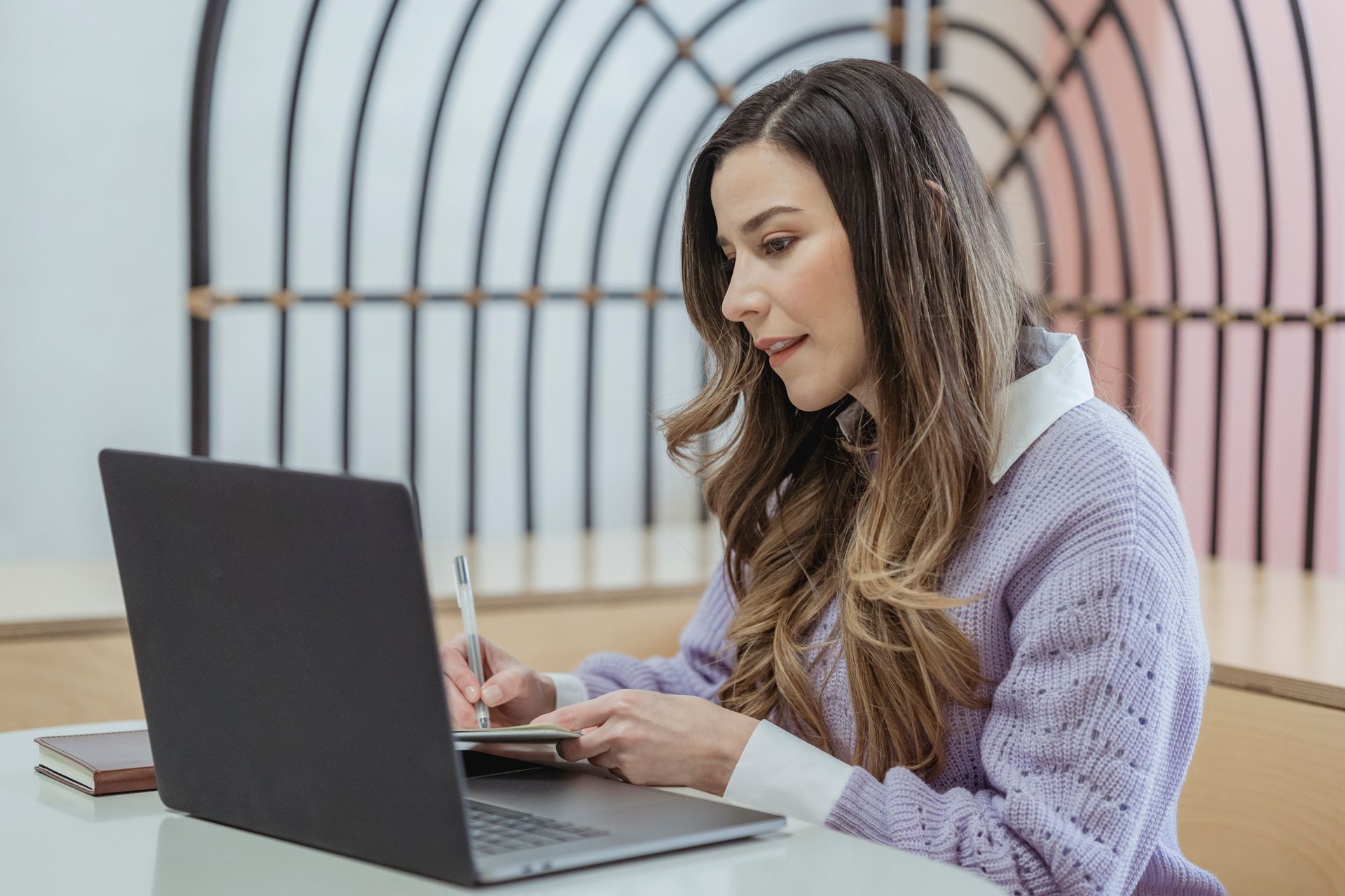 Positive-looking woman in a cardigan and blouse writing notes at a laptop.
